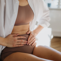 A person sits with hands on their stomach, wearing a white shirt and brown undergarments, suggesting discomfort or pain. The background is softly lit and blurred.