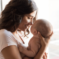 A woman with dark hair smiles warmly at a baby she is holding close. The baby looks back at her. They are near a bright window, creating a soft and gentle atmosphere.