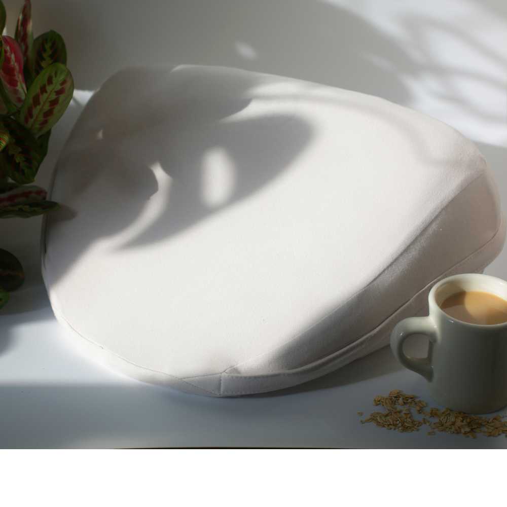 The Dame Pillo - Sex Pillow Wedge in light beige rests on a white surface beside a plant adorned with red and green leaves. Nearby, there's a mug of coffee and scattered oats, with sunlight softly casting shadows that accentuate the product's smooth medical-grade silicone texture.