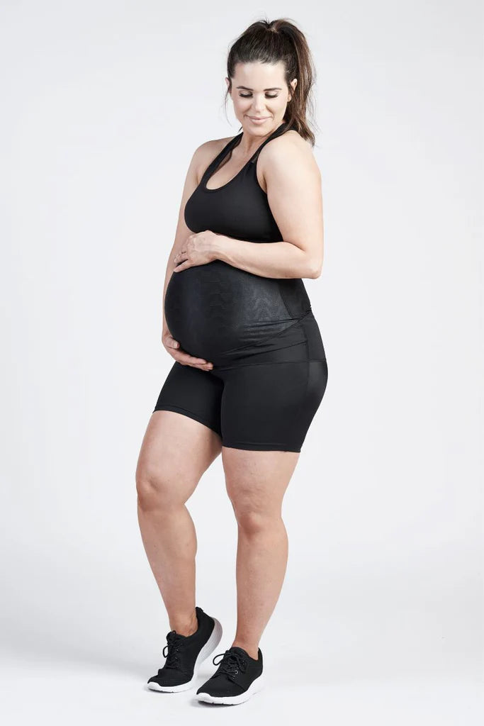 A pregnant woman in athletic wear stands with her legs slightly apart. She is wearing a black tank top and SRC Pregnancy Shorts - Mini Over the Bump by SRC Health. Her hands gently cradle her belly, and she smiles while looking down. With long, dark hair tied back in a ponytail, she seems to be embracing the moment with pelvic floor support for pain relief.