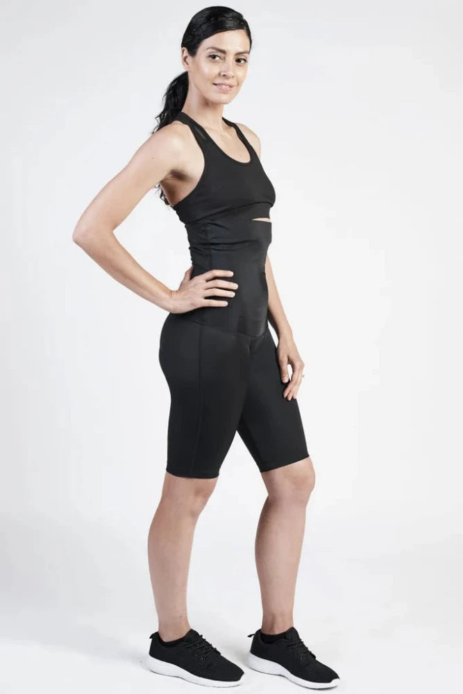 A person stands in a slight profile with one hand on their hip, wearing a black athletic outfit consisting of a sports bra and SRC Postpartum Recovery Shorts from SRC Health, along with black sneakers. The background is plain and well-lit. The person has their hair tied back, exemplifying medical-grade compression apparel ideal for postpartum support.