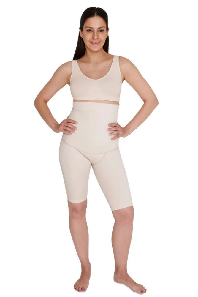 A person with long dark hair stands barefoot against a white background. They are wearing SRC Health's beige medical-grade compression shapewear, including a longline bra and SRC Postpartum Recovery Shorts that are high-waisted and mid-thigh length. The individual has their hands on their hips and is smiling confidently at the camera.