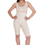 A person with long dark hair stands barefoot against a white background. They are wearing SRC Health's beige medical-grade compression shapewear, including a longline bra and SRC Postpartum Recovery Shorts that are high-waisted and mid-thigh length. The individual has their hands on their hips and is smiling confidently at the camera.