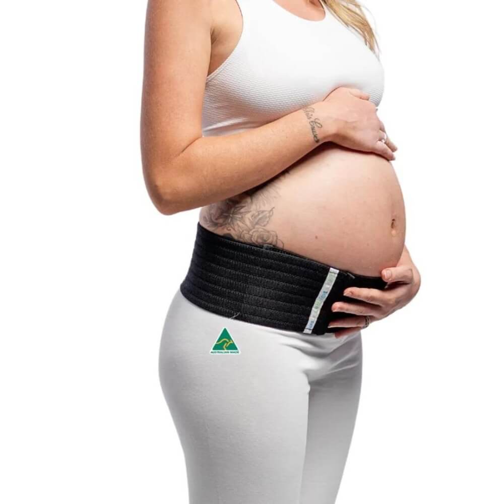 A pregnant woman in a white tank top and pants wears the Belly Bands Maternity Sacroiliac Pelvic Support Belt. She holds her belly, revealing a tattoo on her arm, while the belt offers relief from pelvic girdle pain. The background is white.