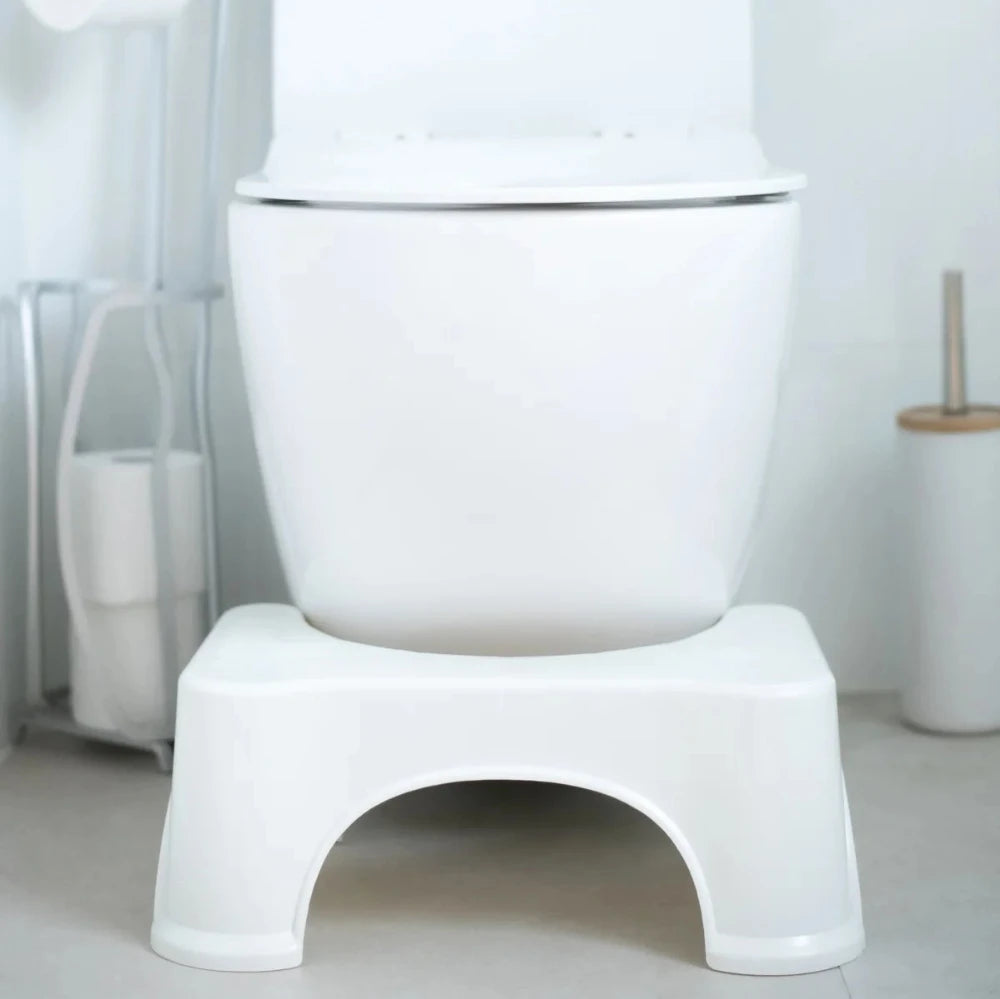 A white toilet paired with the BIEN Toilet Stool from BIEN Australia is positioned at its base to encourage a natural bowel movement. A white toilet brush and holder set, along with a toilet paper holder, contribute to the clean, minimalist decor designed to naturally alleviate constipation.