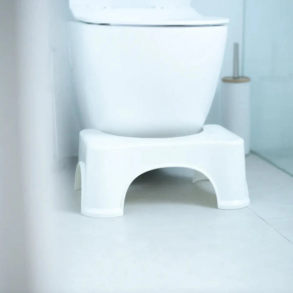 A white bathroom features a modern toilet and a BIEN Toilet Stool by BIEN Australia, which is ergonomically designed to help with proper posture and promote natural bowel movements. A toilet brush in a holder stands next to the toilet.