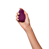 A hand holding the Dame Pom - External Flexible Vibrator, a small, egg-shaped device in a vibrant purple color made from medical-grade silicone, against a white background. The smooth and ergonomic design of the Dame Pom ensures it fits comfortably in the palm of your hand.