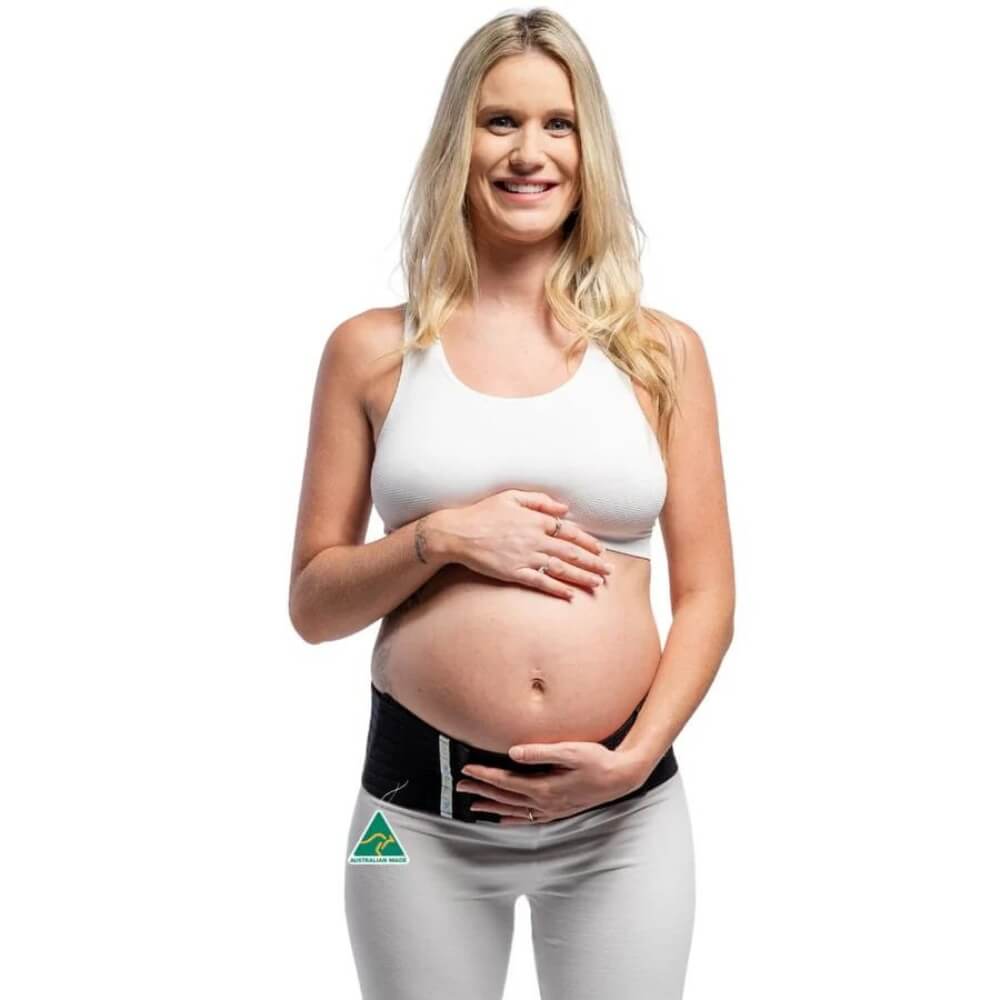 A smiling pregnant woman with long blonde hair, wearing a white tank top and the Belly Bands Maternity Sacroiliac Pelvic Support Belt, stands against a white background. She cradles her belly gently, enjoying the comfort and relief from pelvic girdle pain provided by her Belly Bands support belt.