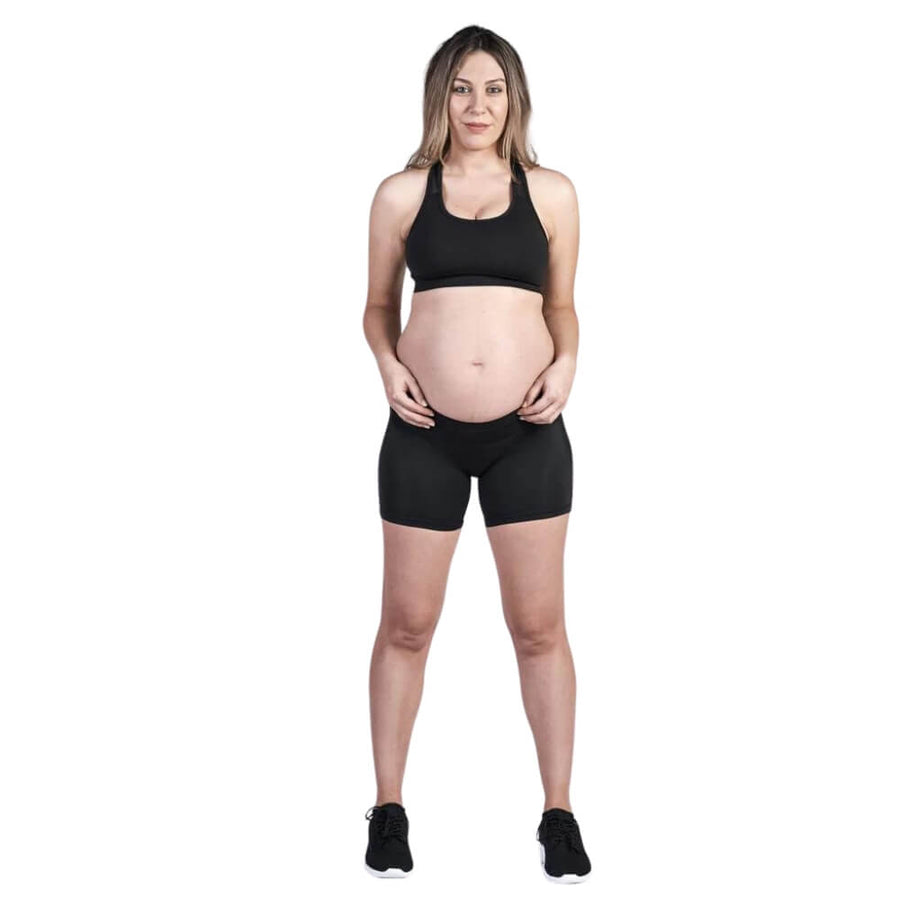 A person with long hair is standing and facing the camera, dressed in a black sports bra, SRC Maternity Shorts Mini by SRC Health, and black shoes. Their hands rest on their hips in a relaxed stance against a plain white background.