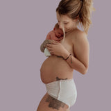 A woman with tattoos, wearing Bubba Bump's Disposable Postpartum Underwear, cradles a sleeping newborn baby against her chest. She gazes at the baby lovingly, showcasing a tender moment of motherhood. The background is a plain, solid color.