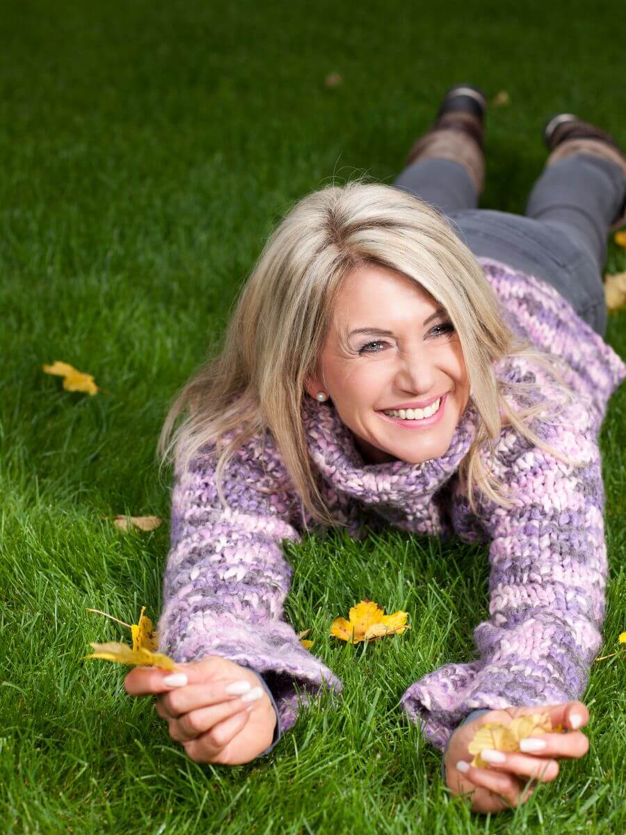 A woman with blonde hair is lying on her stomach on a grassy field, wearing a purple sweater. She is smiling and holding yellow leaves in her hands, surrounded by scattered autumn leaves.