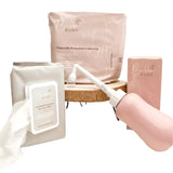 The Bubba Bump Peri Care Bundle is displayed on a wooden stand, showcasing postpartum recovery essentials: distinctive disposable underwear, calming Witch Hazel wipes, and a multi-functional peri bottle. These items are tastefully wrapped in a soft pink package, accented with a small sprig of dried flowers for decoration.