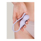 A hand holds a lilac, egg-shaped Perifit Care+ Kegel exercise device next to a person in beige ribbed underwear. It's designed for pelvic floor strengthening with a loop handle for easy use and incorporates biofeedback technology for effective results.