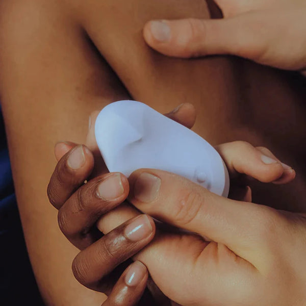 A close-up showcases two hands gently holding the Dame Pom, an external flexible vibrator made from medical-grade silicone, against a person's skin. The softly blurred background accentuates the ergonomic design and interaction between the hands and the Dame product.