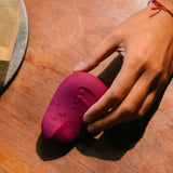 A hand reaches to pick up a pink Dame Pom external flexible vibrator, crafted from medical-grade silicone and adorned with water droplets as it rests on a wooden table. Nearby, a wax-like block sits on a circular tray.