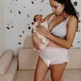 A woman stands in a living room holding a sleeping newborn baby wrapped in a blanket. She is wearing a grey tank top and Bubba Bump Disposable Postpartum Underwear. The background features a light-colored wall with a scattered pattern and a beige couch.