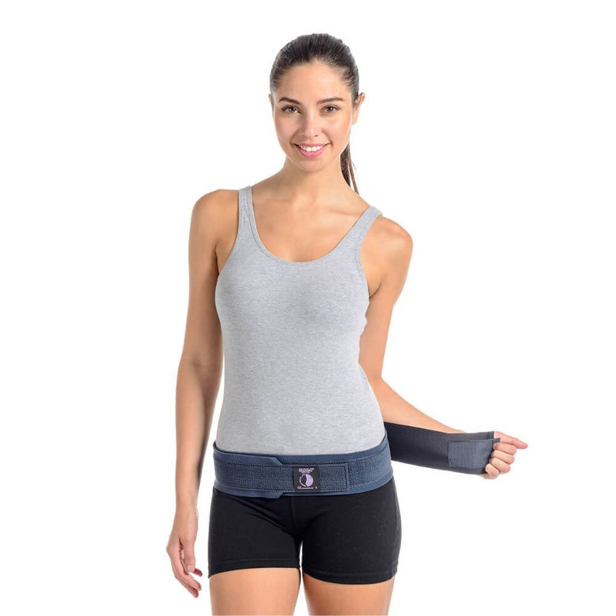 A person in a gray tank top and black shorts is smiling as they fasten the Serola Belt - Pregnancy Sacroiliac Belt around their waist, offering pelvic support. The background is plain white.