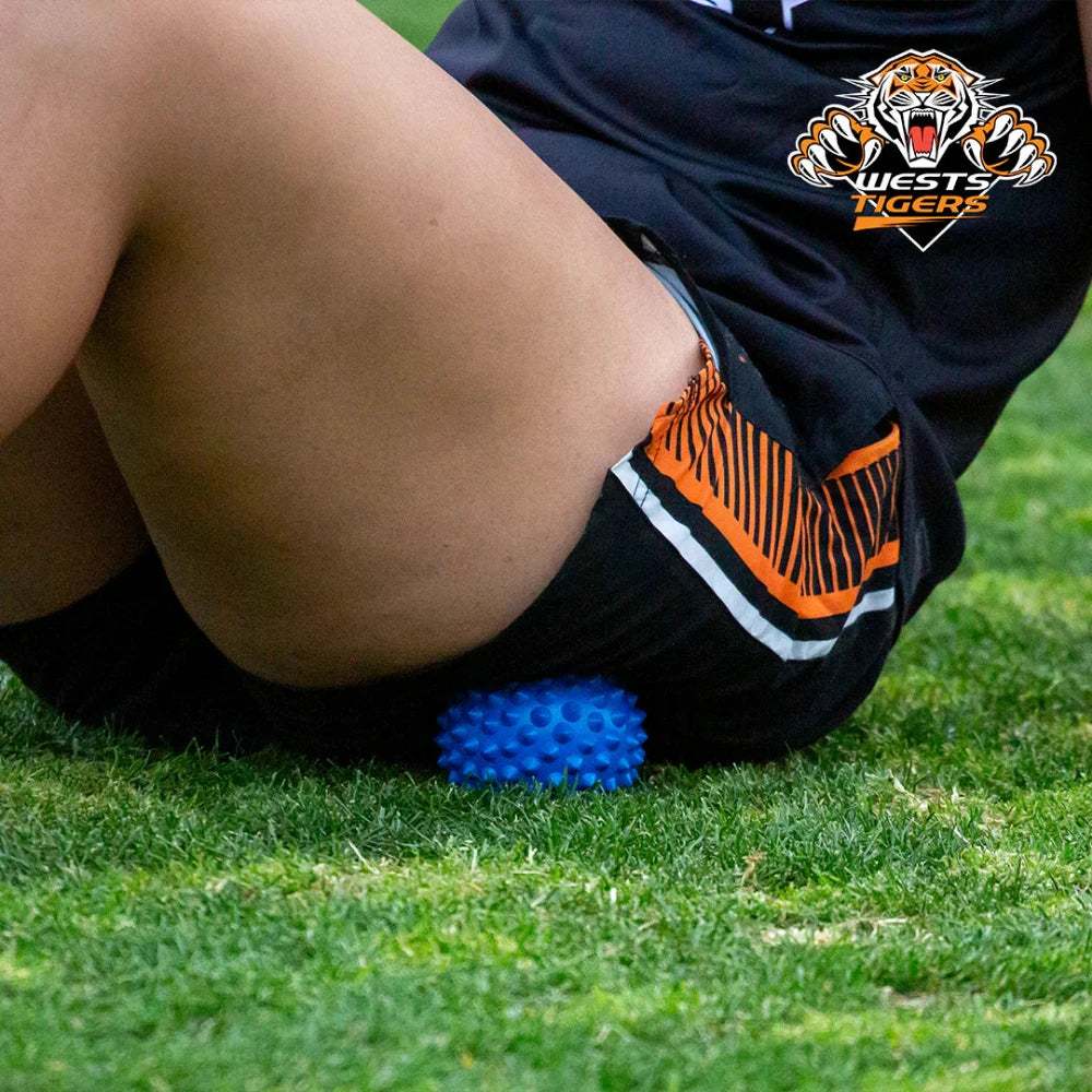 A person with the Wests Tigers team logo on their shorts is using a blue spikey massage ball on their leg.