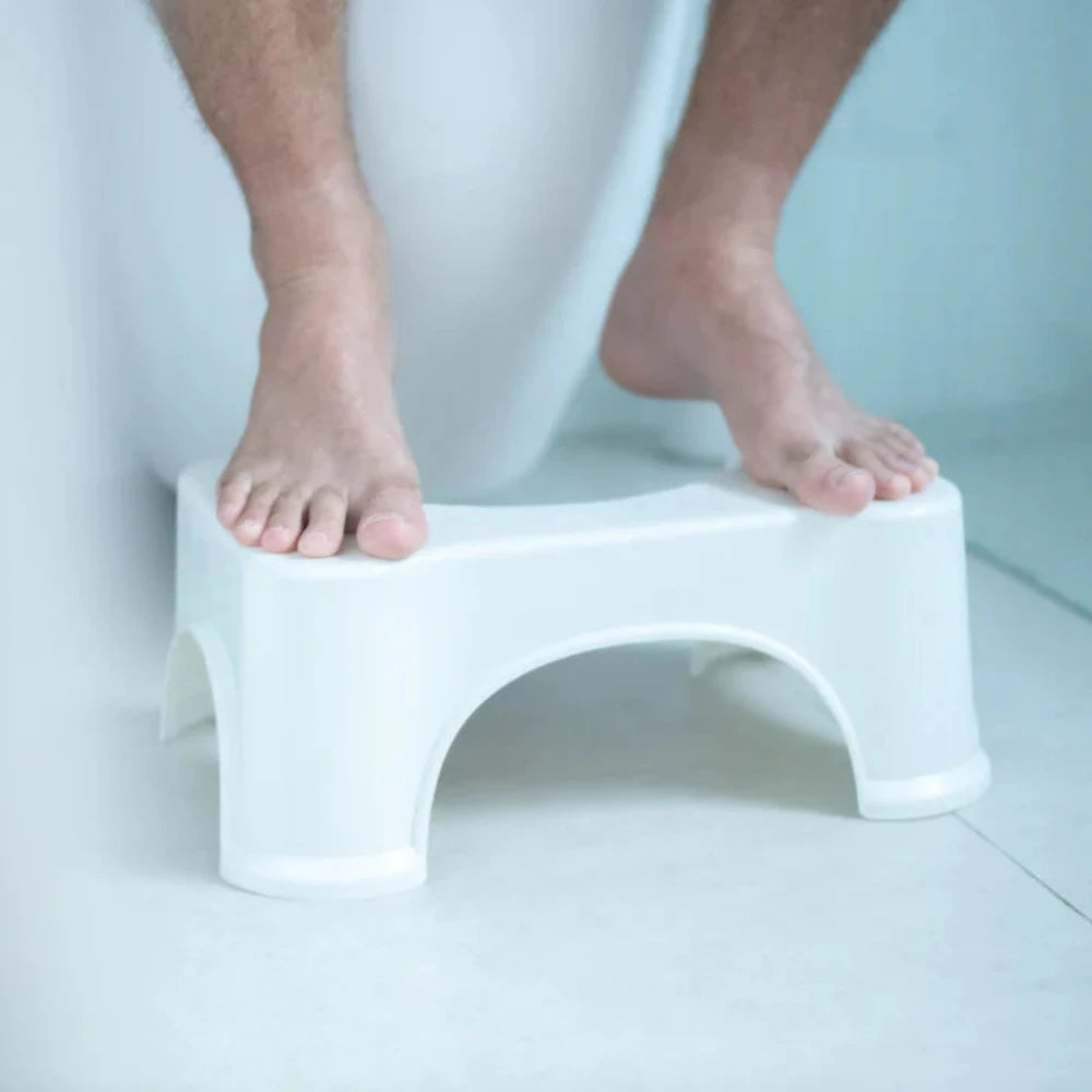 A person stands with both feet on a BIEN Toilet Stool by BIEN Australia against a light-colored background. The white plastic stool, designed to alleviate constipation, features curved arches on its sides and rests on a smooth surface. The lower legs and feet are visible.